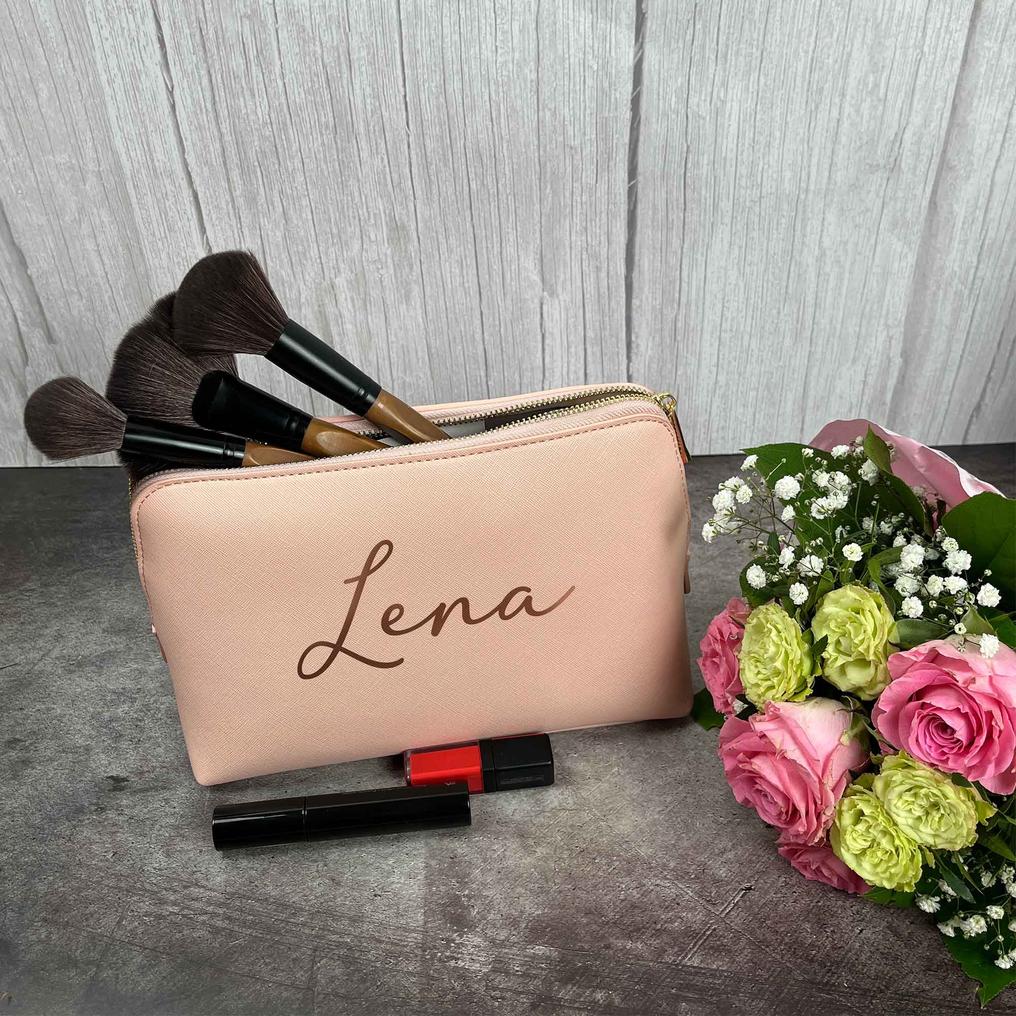 Rosa Kosmetiktasche mit Namen Lena, gefüllt mit Make-up Pinseln und Blumenstrauß daneben