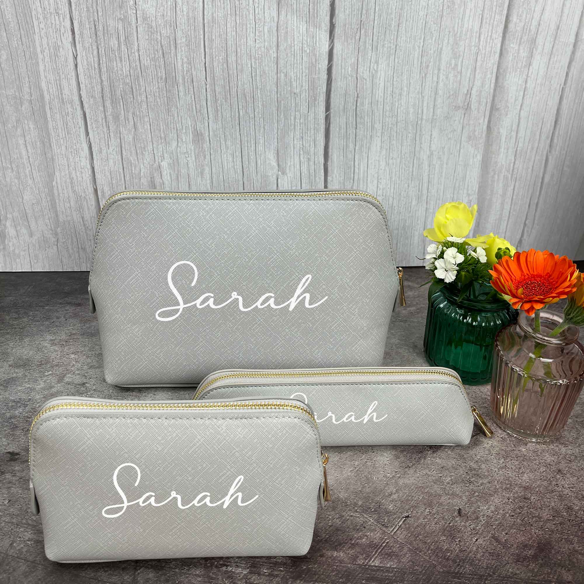 Drei personalisierte Kosmetiktaschen in Grau mit weißem Namen und Blumen im Hintergrund