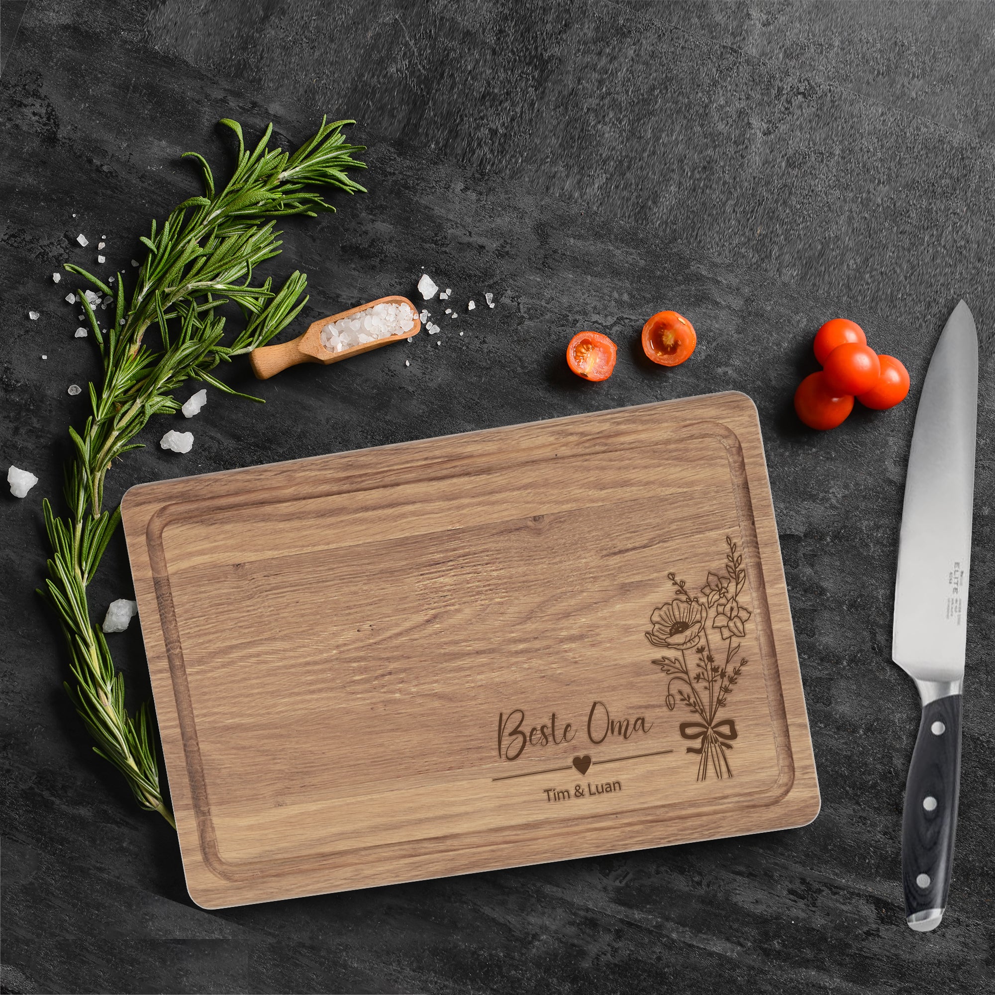 Personalisiertes Schneidebrett „Beste Oma“ auf dunkler Arbeitsplatte mit Messer, Rosmarin und Tomaten