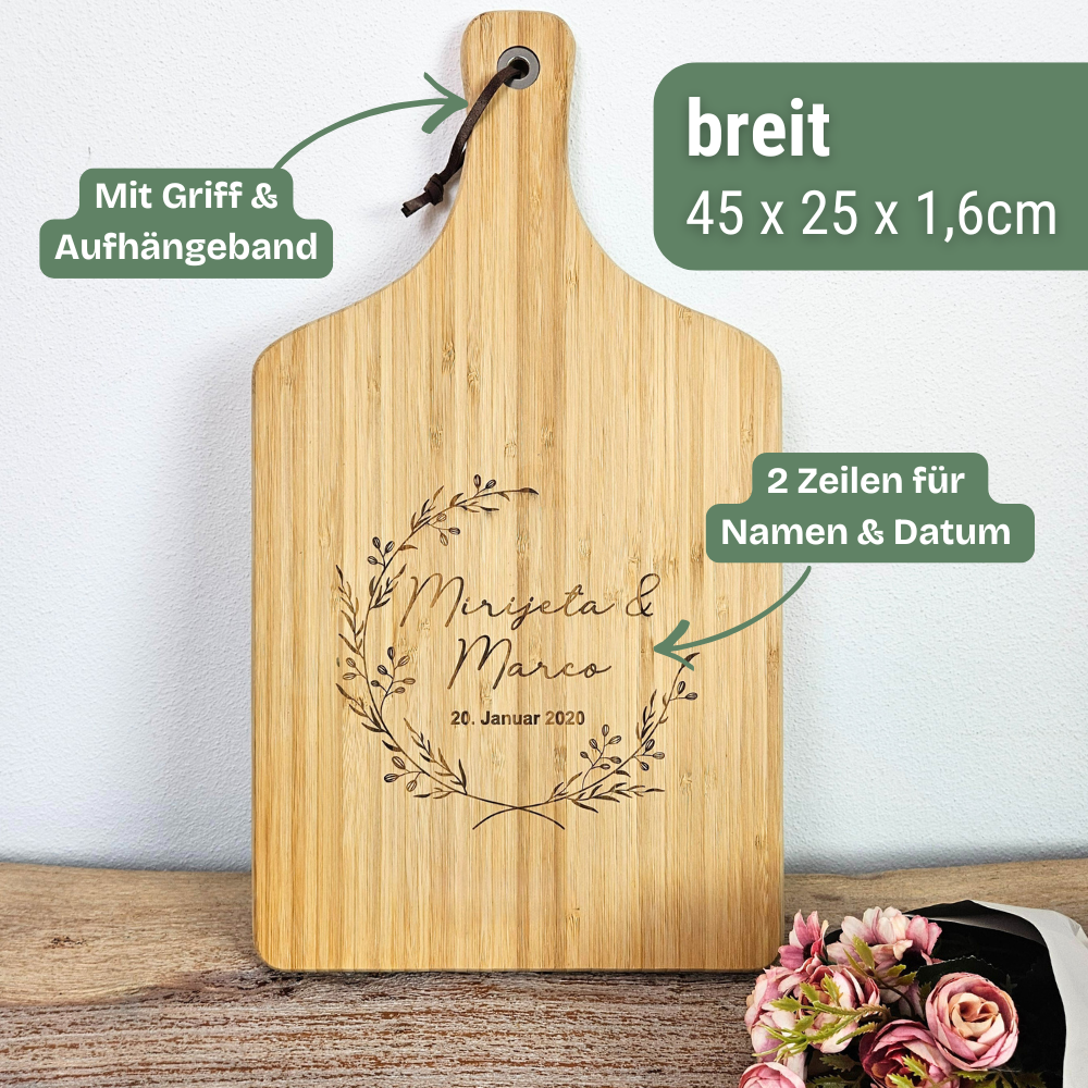 Bambus Schneidebrett personalisiert zur Hochzeit, dekorativ inszeniert mit Blumen im Hintergrund. Geschenk zur Hochzeit mit Gravur von Namen und Datum für Paare.